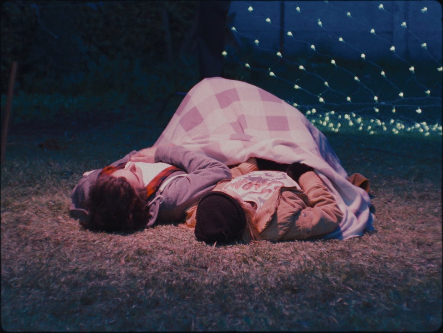Two people lying on the grass at night, covered with a checkered blanket, under hanging lights